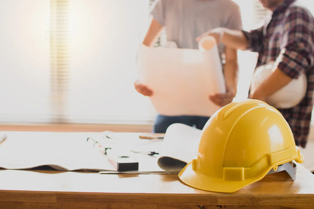 A yellow hard hat on a desk beside rolled drawings, with two people reviewing plans